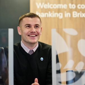 Image of a male Cash Access UK employee at the service counter. Looking up and smiling, while wearing a branded pin badge.