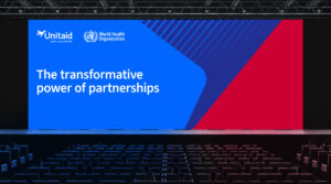 A conference stage with a large screen displaying the text “The transformative power of partnerships” and the logos of Unitaid and the World Health Organization.